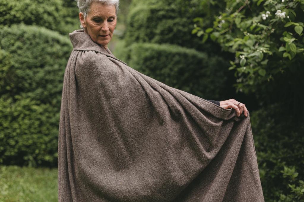 Older white woman with gray hair standing surrounded by greenery wearing 207 Kinsale Cloak without hood holding the cloak out with one arm to show length.