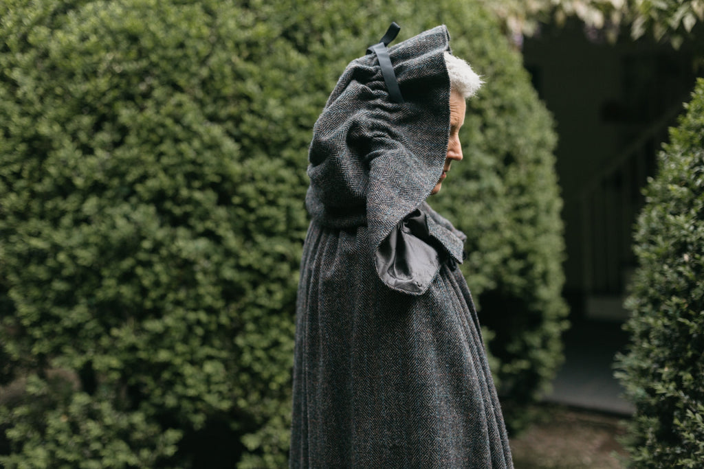 Close up of older white woman standing surrounded by greenery wearing 207 Kinsale Cloak with hood up.