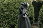 Close up of older white woman standing surrounded by greenery wearing 207 Kinsale Cloak with hood up.